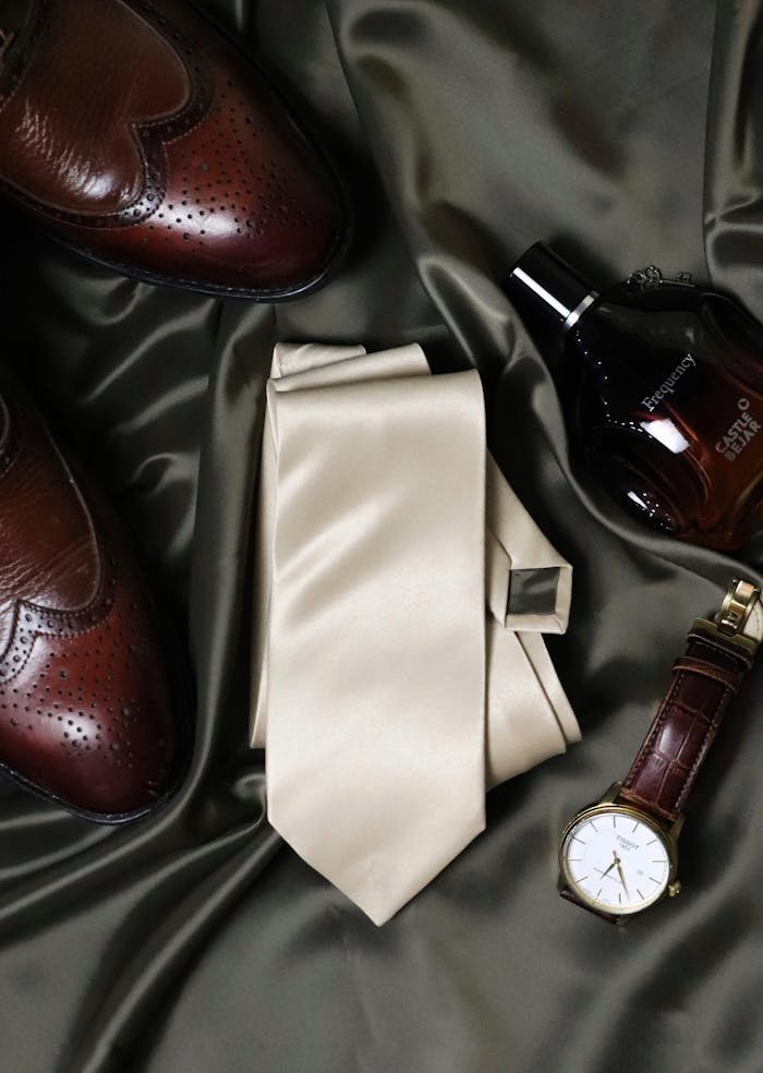 A flat lay of elegant mens accessories including a necktie, leather shoes, cologne, and a watch on a satin fabric.
